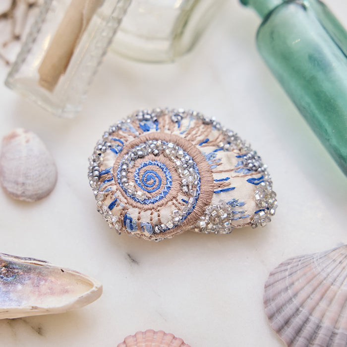 Craftspring’s Moon Snail Shell Ornament — made of satin with intricate beaded and embroidered details — displayed among seashells and glass bottles, creating a festive coastal holiday scene, ethically crafted by artisans.