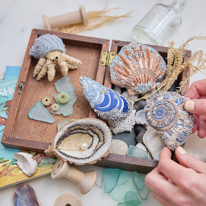 Craftspring’s Mussel Shell, Oyster, Moon Snail Shell, Scallop Shell, and Hermit Crab ornaments — all intricately beaded and embroidered, the shells made of satin and the Oyster and Hermit Crab made of felt — displayed in a box of ocean treasures with seashells and sea glass, creating a playful and festive holiday scene, ethically crafted by artisans.