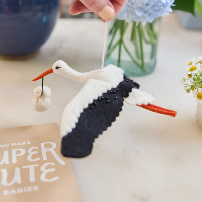 Handmade felt New Baby Stork ornament from Craftspring, featuring a stork carrying a bundle with delicate beaded and embroidered details—displayed hanging surrounded by flowers and a card, perfect for nursery decor and thoughtful baby shower gifting, ethically crafted by artisans using sustainable wool.
