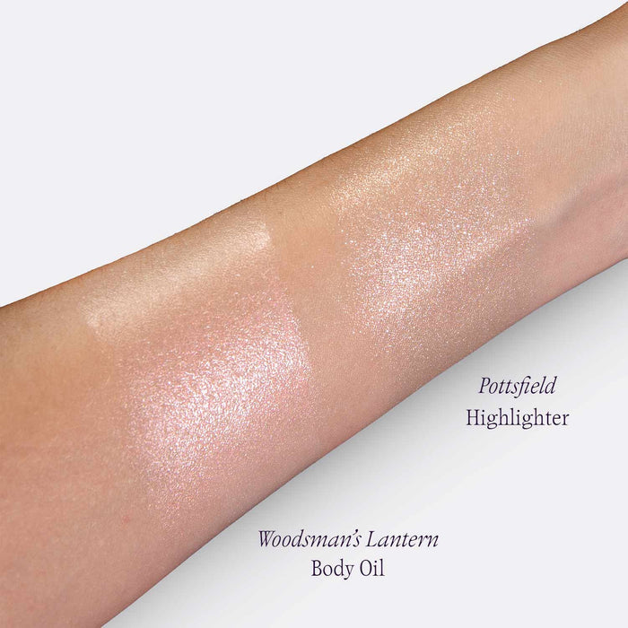 A close-up of a forearm showing two shimmering highlighter swatches on skin, labeled 'Pottsfield Highlighter' and 'Woodsman's Lantern Body Oil'.