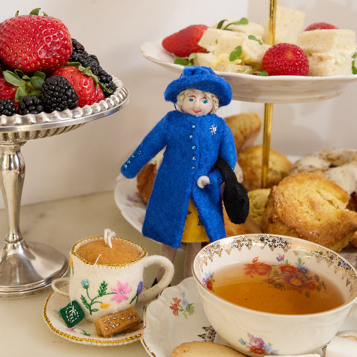 Queen Elizabeth II felt ornament wearing royal blue coat and hat, with black handbag and detailed embroidery. Handmade wool Christmas ornament honoring British royalty. Fair trade and ready to hang. Pictured with afternoon tea foods and a felt tea cup