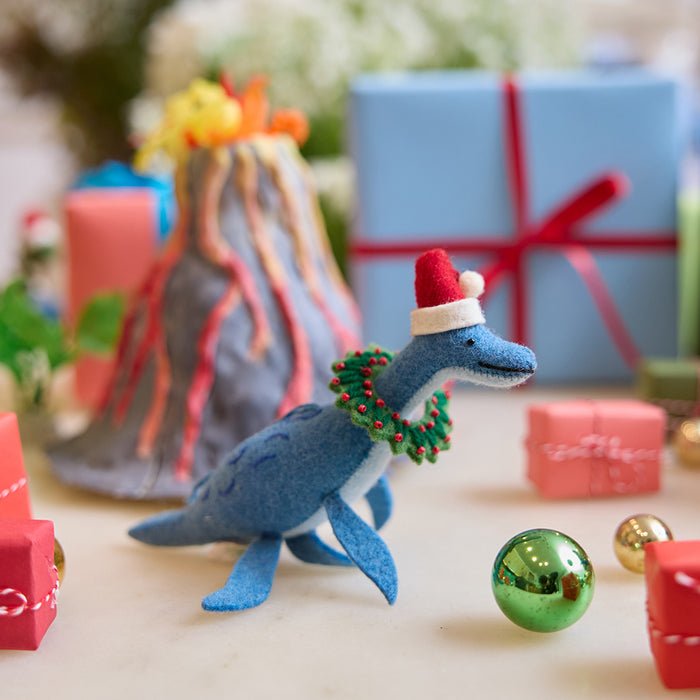 Handmade felt Santa Plesiosaurus ornament from Craftspring, featuring a dinosaur with a Santa hat, festive wreath, and beaded and embroidered details—displayed with a volcano school project and wrapped presents, perfect for whimsical Christmas gifting and imaginative holiday decor, ethically crafted by artisans using sustainable wool.