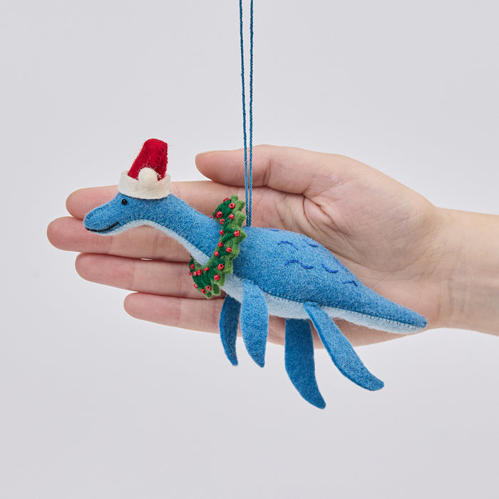Handmade felt Santa Plesiosaurus ornament from Craftspring held in front of a hand to show size—featuring a dinosaur with a Santa hat, festive wreath, and intricate beaded and embroidered details, ethically crafted by artisans using sustainable wool.