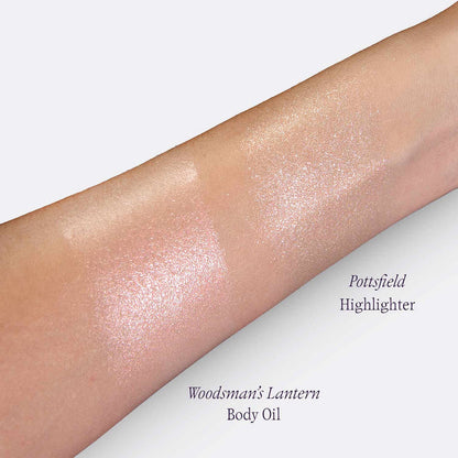 A close-up of a forearm showing two shimmering highlighter swatches on skin, labeled 'Pottsfield Highlighter' and 'Woodsman's Lantern Body Oil'.